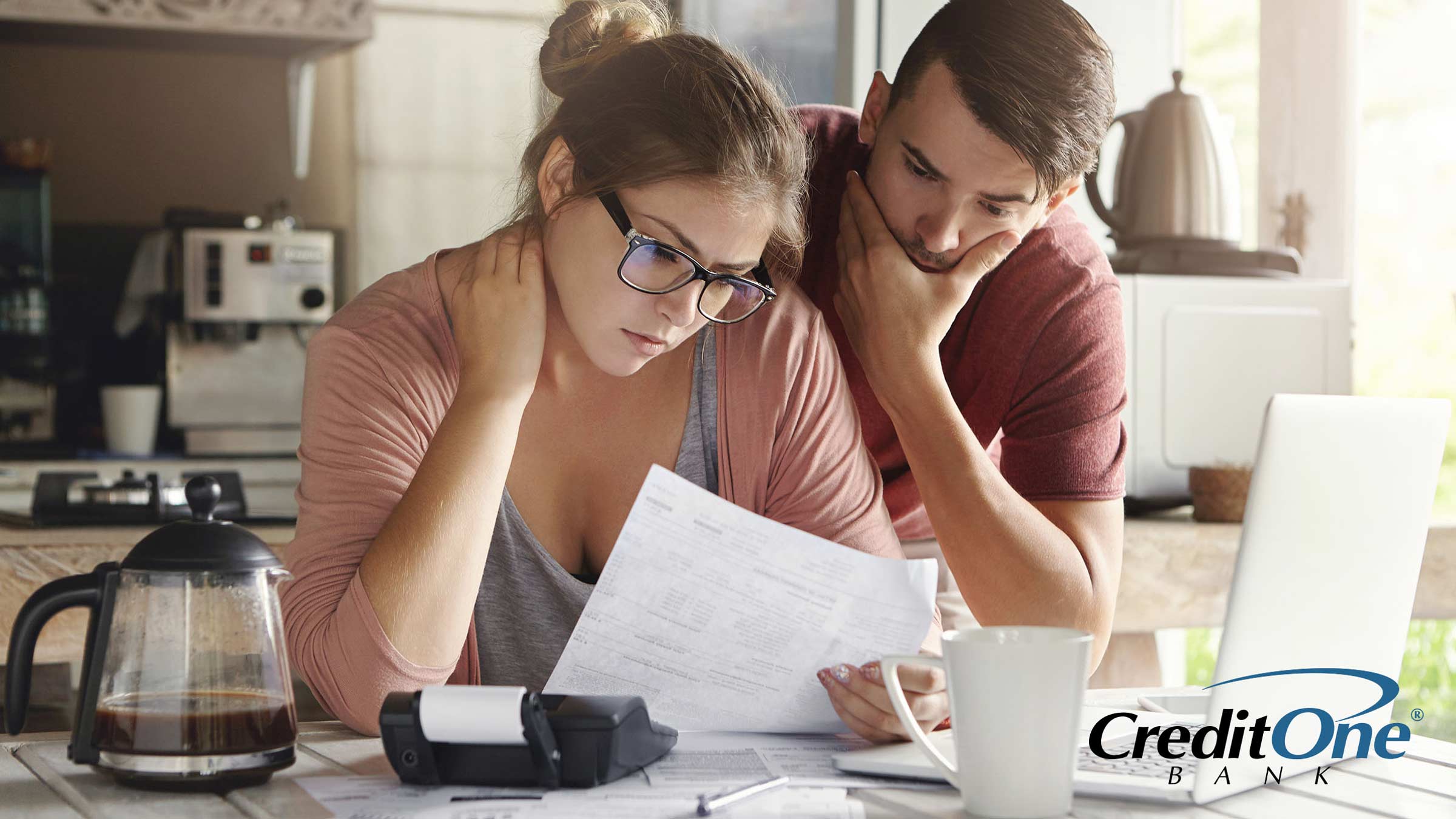 A concerned couple reviews a financial statement at their kitchen table, which has several documents, a calculator, coffee pot and laptop on it. Perhaps they forgot a bill and are considering what happens when you make a late credit card payment.
